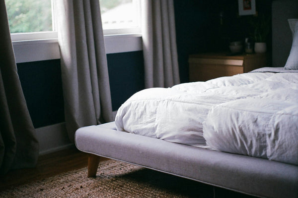 A neatly made white bed beside a bright window with daylight coming through in a calm bedroom