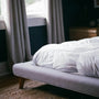 A neatly made white bed beside a bright window with daylight coming through in a calm bedroom