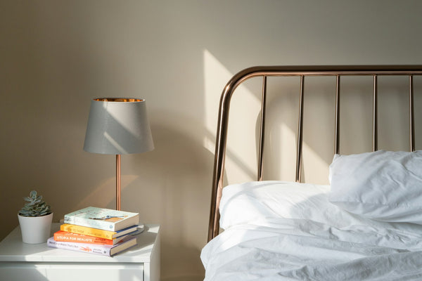 Warm morning sunlight falling across a cosy bedroom with a brass bed, bedside lamp, plant and books