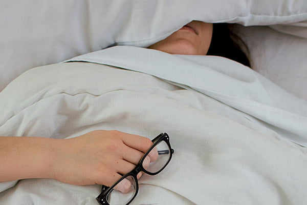 A person lying in bed with a pillow pressed over their face, glasses in hand