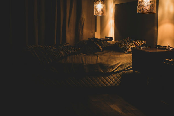 A dimly lit, cosy bedroom at night with warm lamp glow and soft white linen — the calm setting of a sleepmaxxing routine.