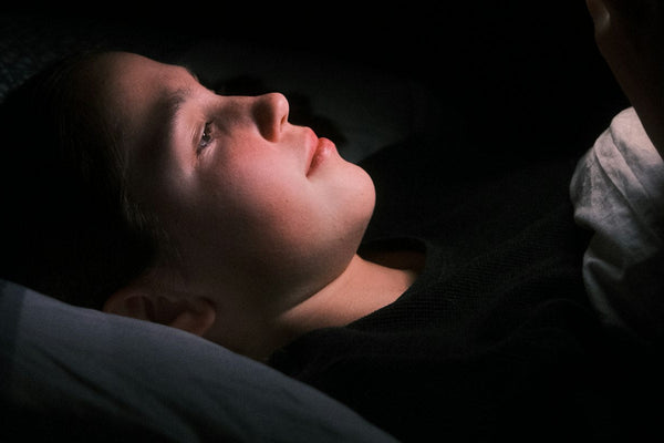 A woman lying in bed in a dark room, her face lit by the glow of a phone screen