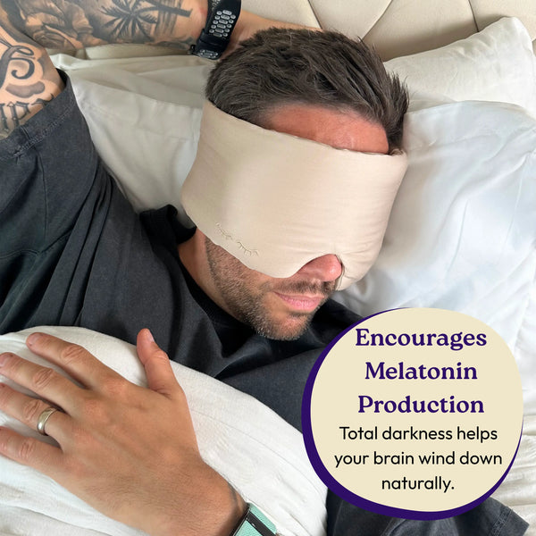 Man lying in bed with a sleep mask on, surrounded by pillows.